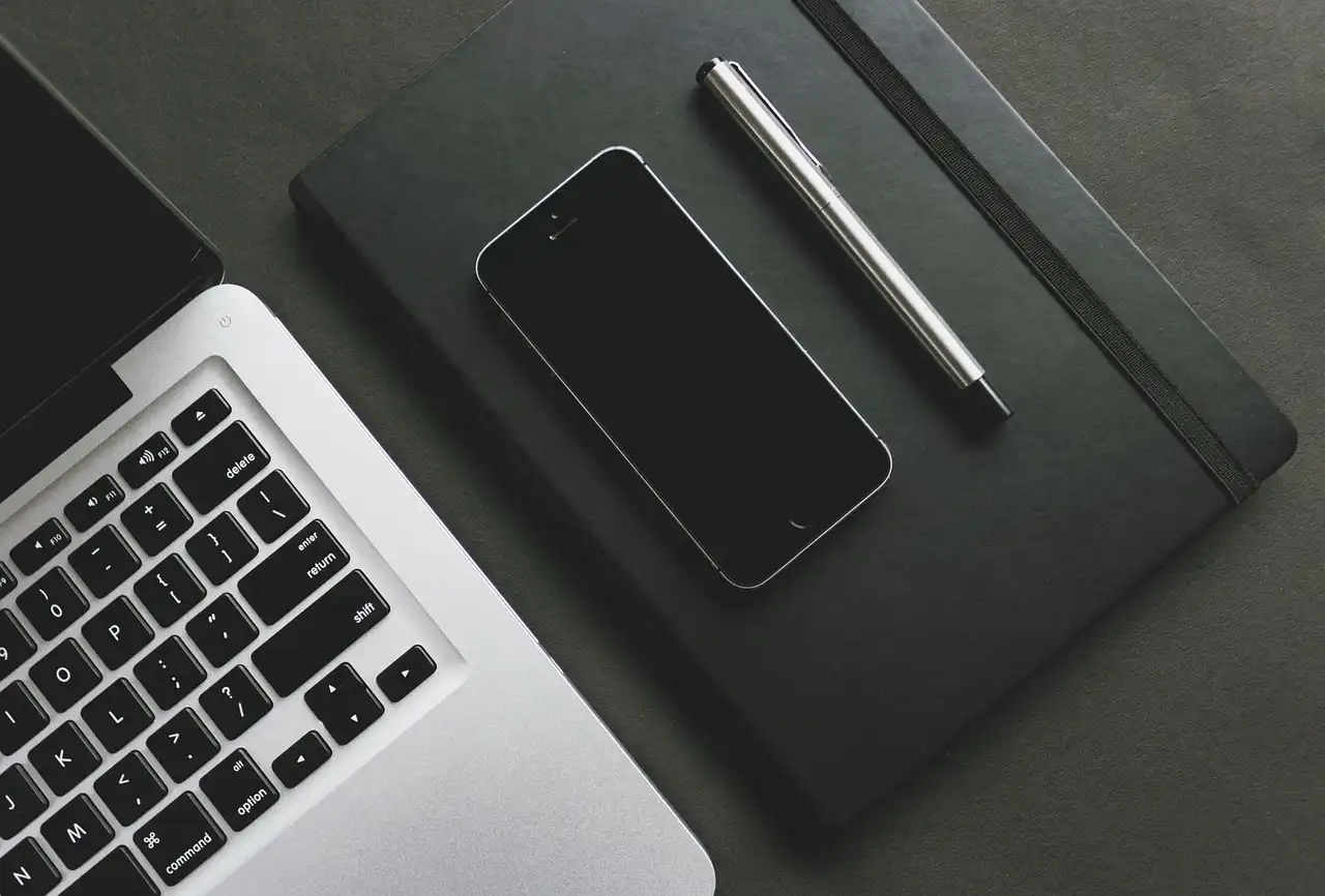 A top-down view of a silver laptop, a black smartphone, a black notebook, and a silver pen on a dark table surface."