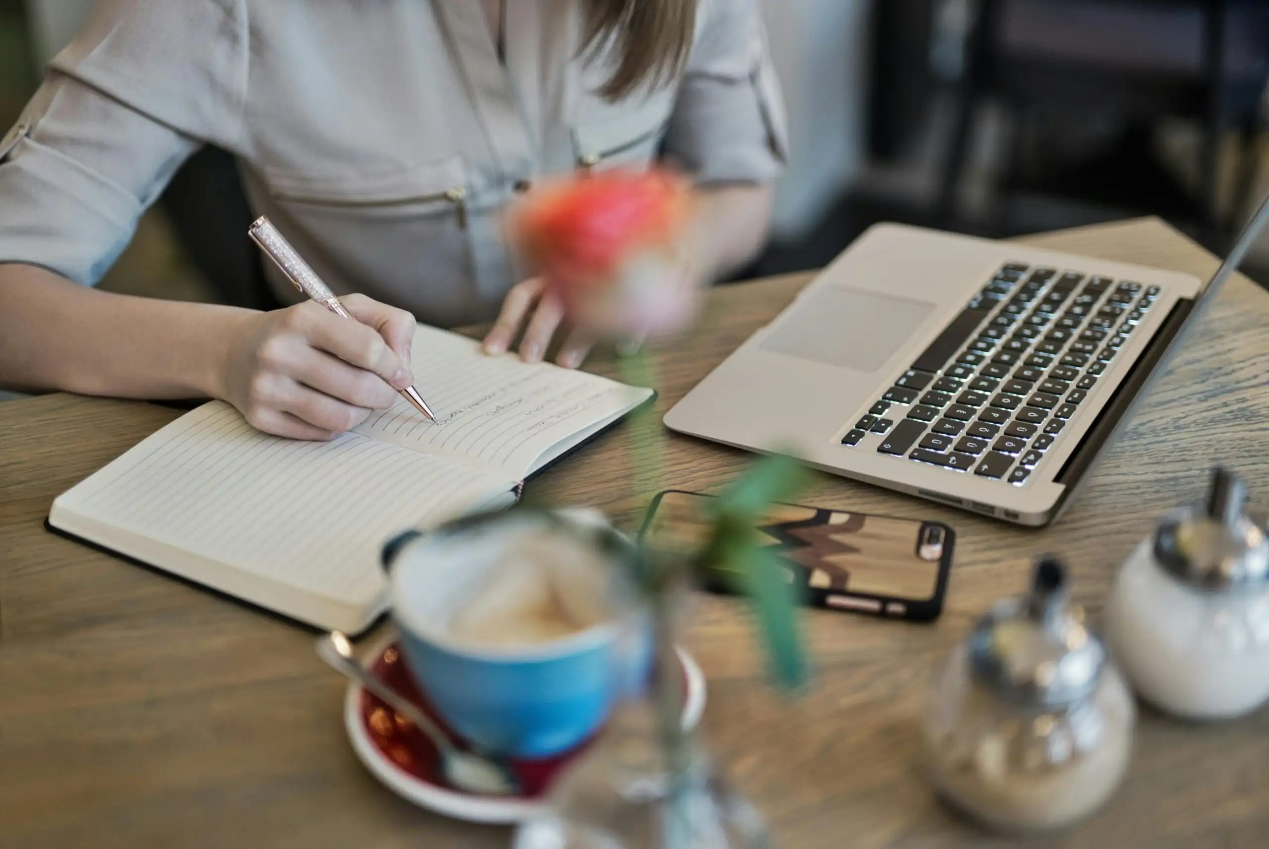 A woman writing on a book, a laptop by side representing Content writing